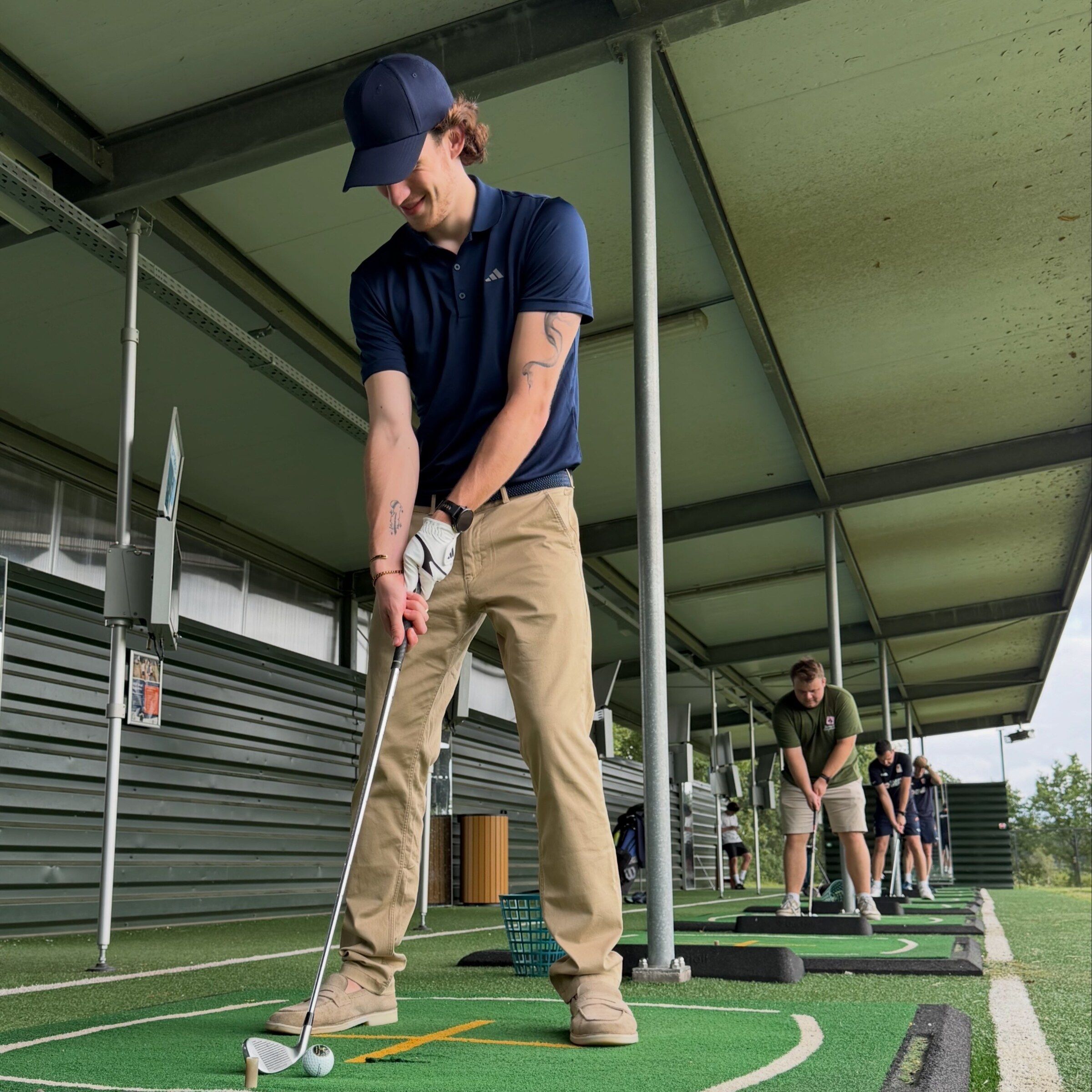 Sven met pet en golfclub boven de bal op een overdekte afslagbaan, met een bak ballen voor zich